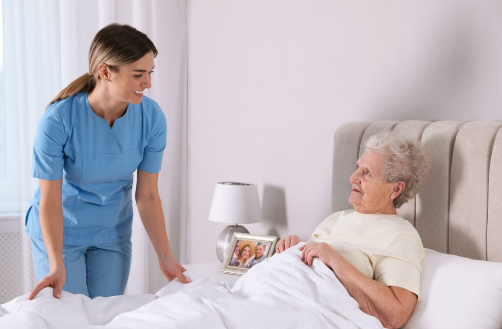 A female caregiver smiling while assisting an older woman who is laying down in bed
