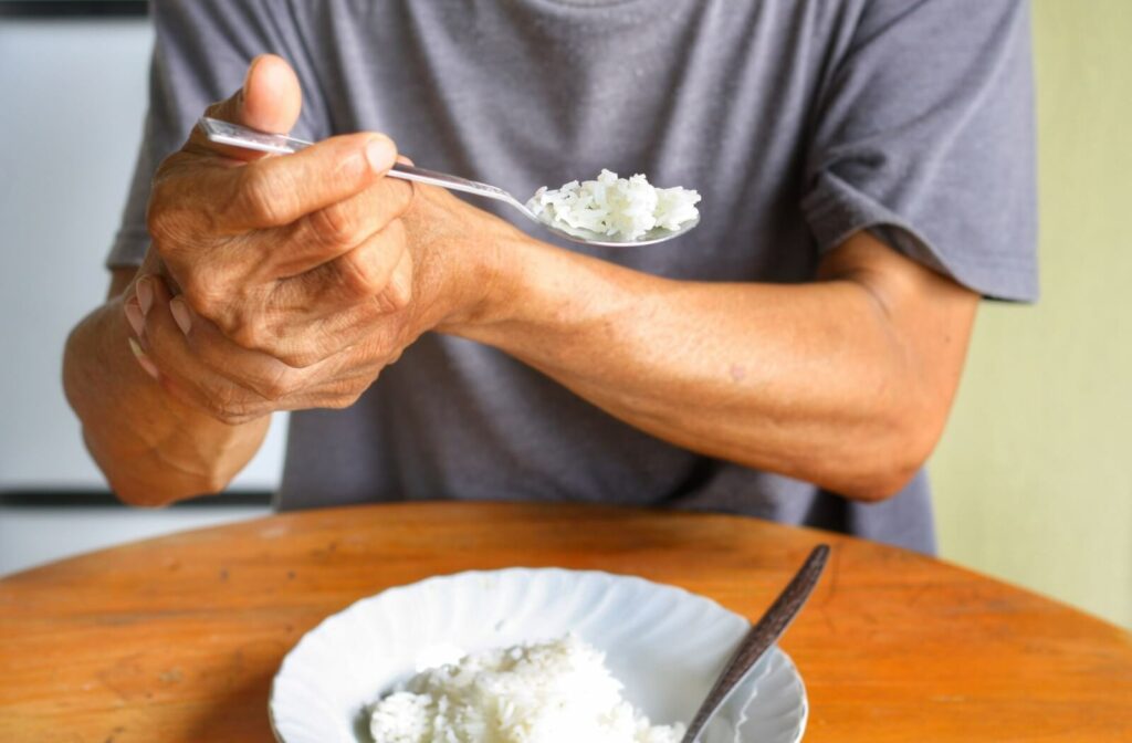 An older adult holding his hand to prevent his tremors spilling the rice from his spoon.