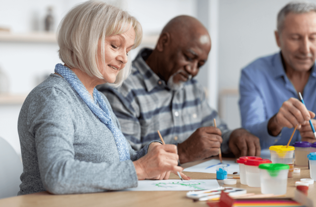 3 seniors sit together happily painting on plain paper in a bright room.