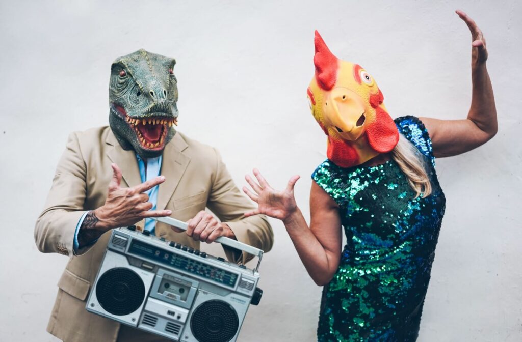 An older couple dressed for Halloween, one in a dinosaur mask holding a boombox, and one in a chicken mask and sequin dress