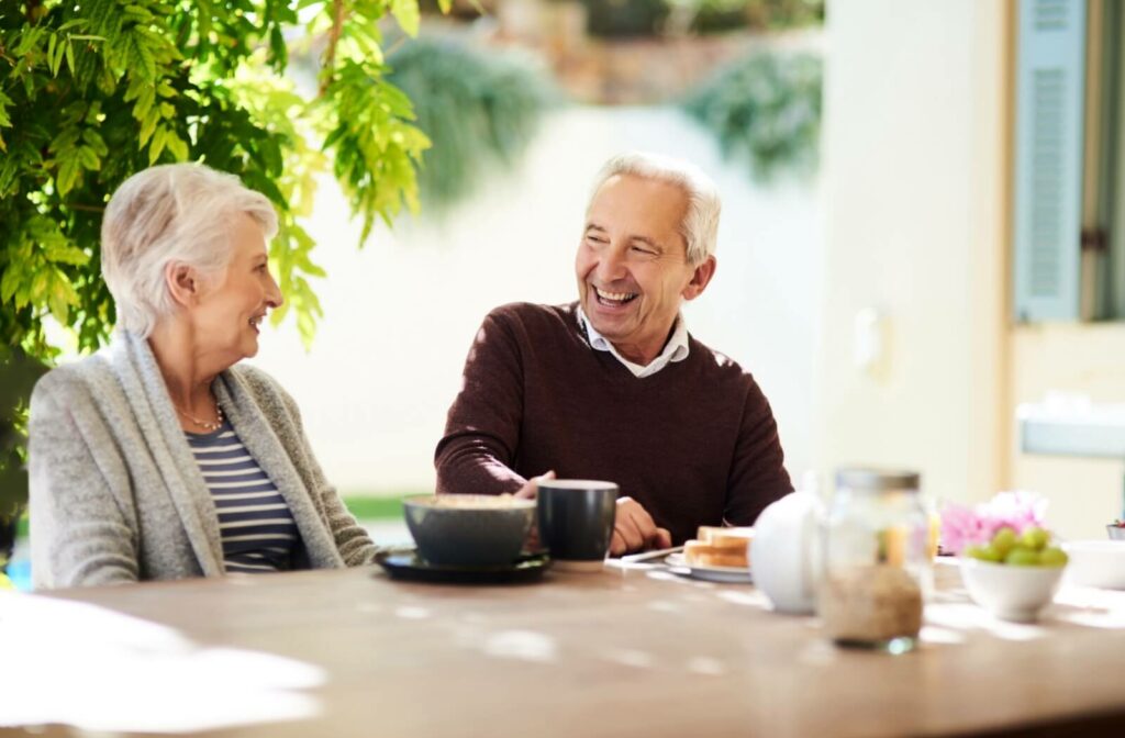 A senior couple laughs with each other while having breakfast and coffee outdoors in the sunshine